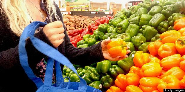 A woman is shopping at a farmers market with a reusable shopping bag. She is looking at colorful bell peppers.