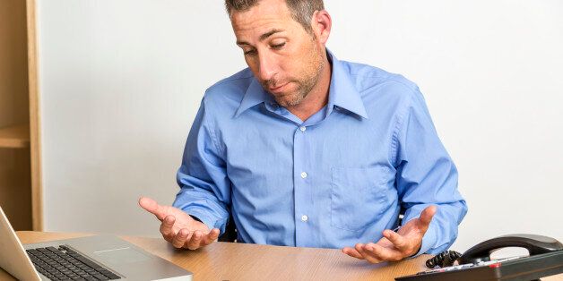 Businessman with his hands out in frustration. He is sitting at a desk surrounded by a laptop, smart phone, tablet computer, and desk phone.