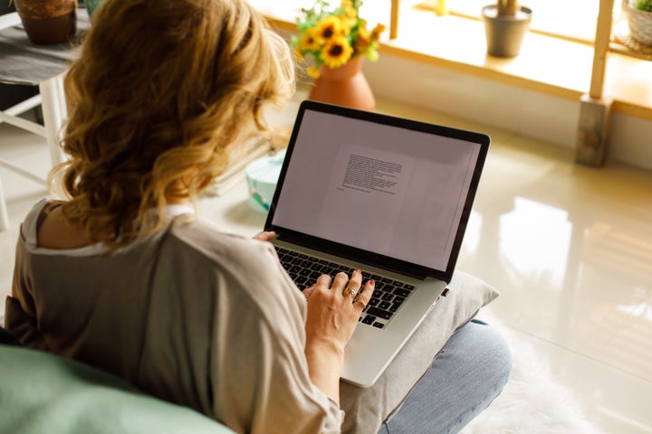 Over the shoulder view of casual mid adult businesswoman sitting on floor in her bedroom and typing data on laptop while working from home.