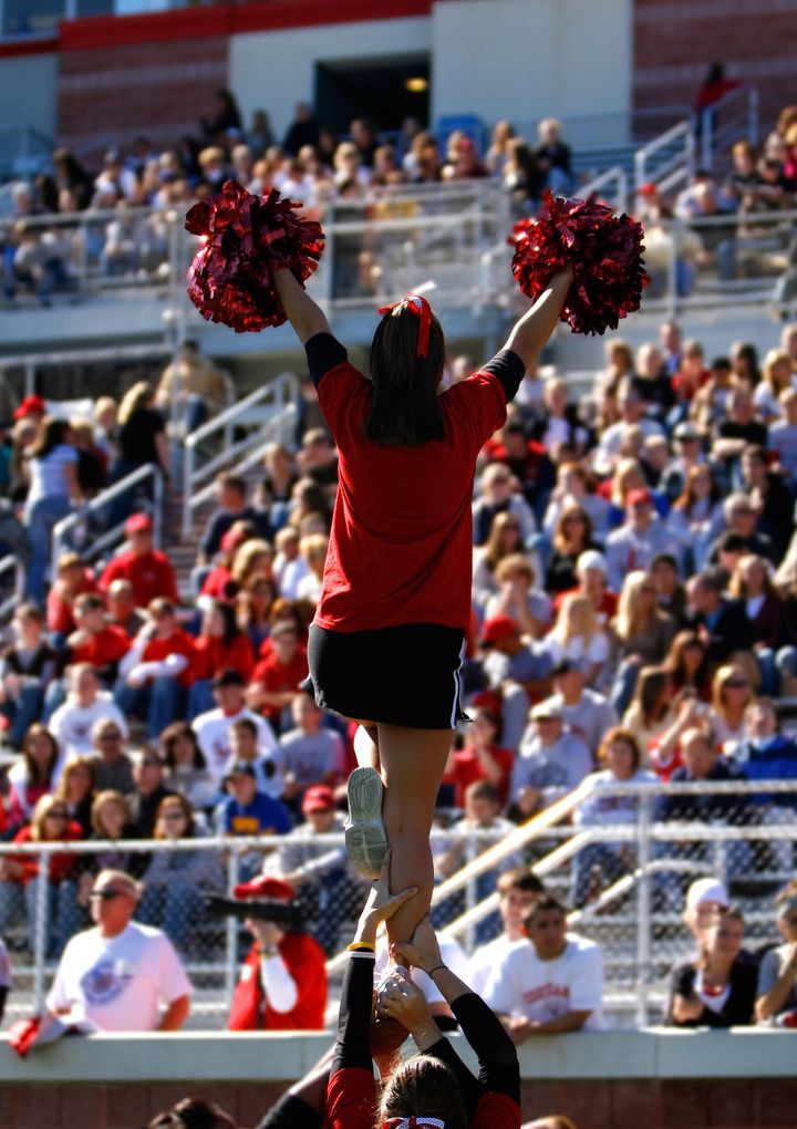 A football cheerleader is raised into the air in front of a large collegiate crowd.
