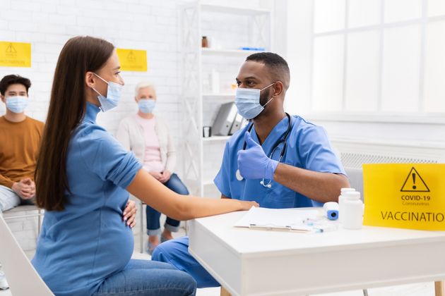 Successful Coronavirus Vaccination. Doctor In Face Mask Gesturing Thumbs-Up To Vaccinated Against Corona Virus Female Patient Sitting In Hospital. Covid Vaccine Injection And Immunization Campaign