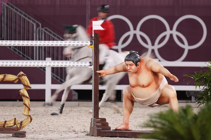 A show jumper rides behind an obstacle with a sumo wrestler beside it in an event at the Tokyo Olympics.