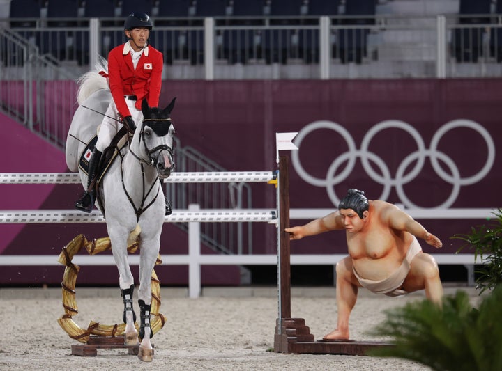 A rider clears the barrier as the statue of a sumo wrestler looks in the Tokyo Olympics equestrian competition. )