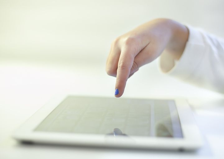 a woman hands with blue nail polish with white tshirt touching the tablet in a ehite room with white table.