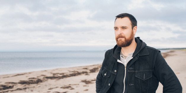 A man with a red beard and denim vest looks down a beach near sunset.