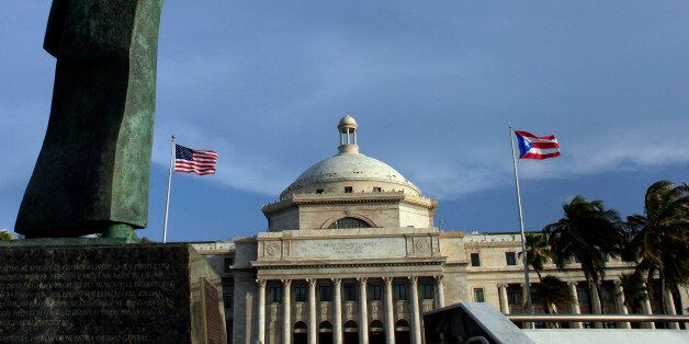 In this Wednesday, July 29, 2015 photo, a bronze statue of San Juan Bautista stands in front of Puerto Ricos Capitol as U.S. and Puerto Rican flags fly in San Juan, Puerto Rico. Mired in an ongoing economic crisis the government has tried to boost revenue by hiking the sales tax to 11.5 percent, higher than any U.S. state, and closing government offices. (AP Photo/Ricardo Arduengo)