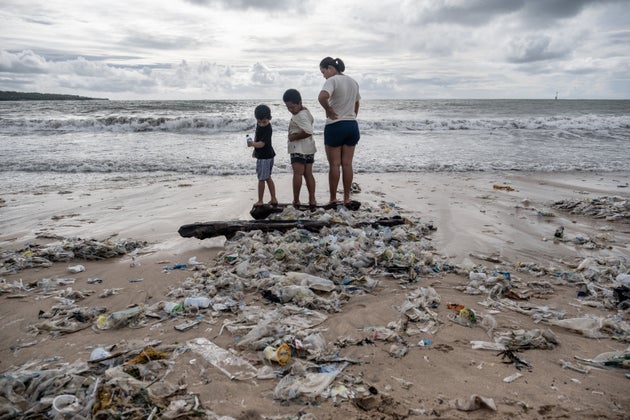 A woman with two boys stand on some wood which is covered by plastic trash at a beach in