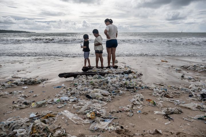 A woman with two boys stand on some wood which is covered by plastic trash at a beach in Bali.