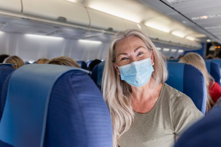 A front-view shot of a mature caucasian woman sitting on an airplane wearing a protective face mask, she is excited to be traveling on holiday.