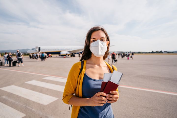 Portrait of young woman with face mask, passport and ticket standing on airfield.