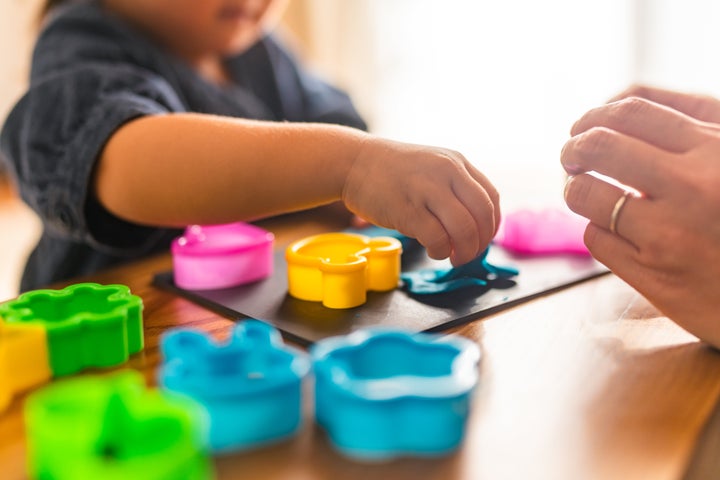 A cute small girl is making shapes using colorful clay with her mother in the living room at home.