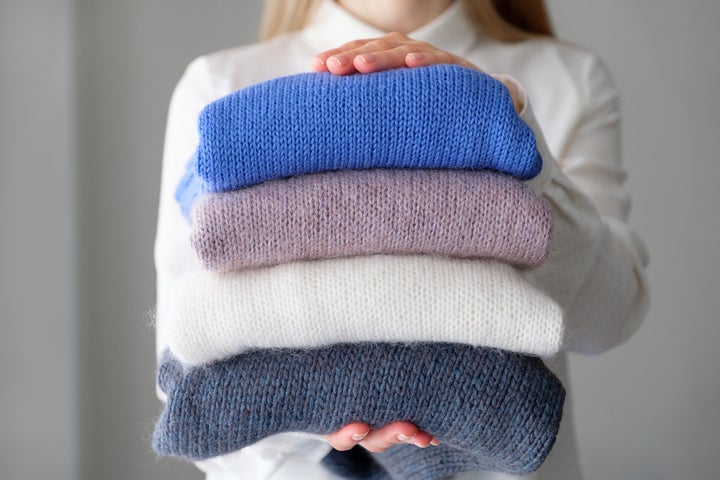 A woman or a girl holds an armful of knitted things of different colors, stacked in a pile, in the room. Winter and autumn warm cozy sweaters for charity. The concept of storage, care and washing of handmade products. A copy of the text space.