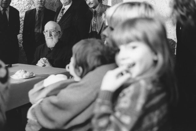 Ο Αββά Πιέρ στο βάθος της φωτογραφίας, περιστοιχισμένος από άντρες με κουστούμια και σε πρώτο πλάνο ένα παιδάκι που κοιτάει την κάμερα και χαμογελάει