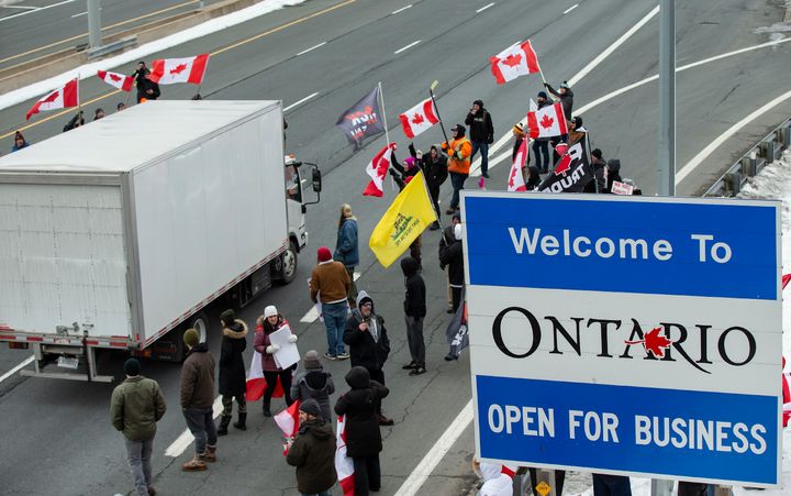 A truck passes Ontario Provincial Police officers and demonstrators in opposition to COVID-19 mandates on the Toronto-bound QEW highway after crossing the Peace Bridge in Fort Erie, Ontario on Saturday, Feb. 12, 2022. (Aaron Lynett /The Canadian Press via AP)