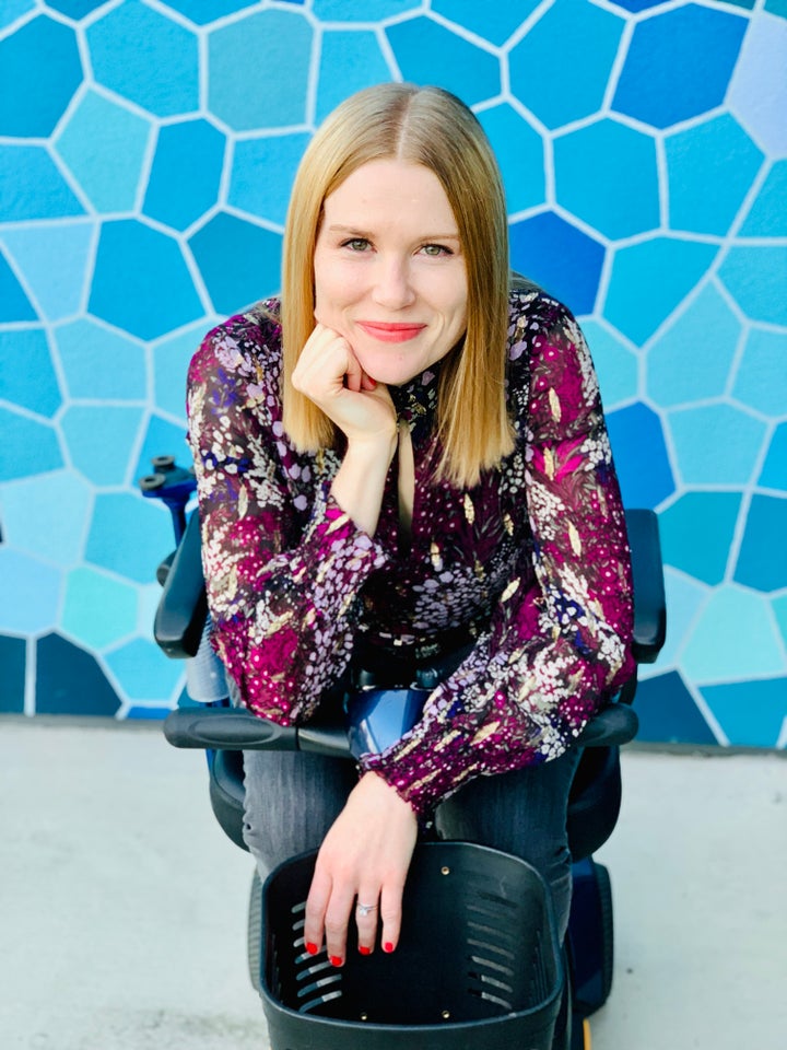 [Image description: A blonde, white woman wearing a burgundy patterned shirt sits on a mobility scooter in front of a blue background with a variety of geometric shapes.]<br><br>"My disabled life is vibrant," the author writes. "My disabled life is a creative force that imagines a new world where we don’t have to endure people being encouraged by my death because I was unwell to begin with."