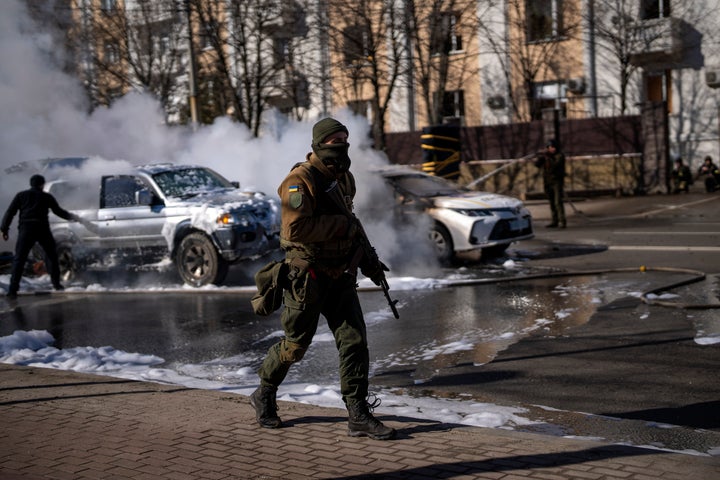 Ukrainian soldiers take positions outside a military facility as two cars burn, in a street in Kyiv, Ukraine, Saturday, Feb. 26, 2022. (AP Photo/Emilio Morenatti)