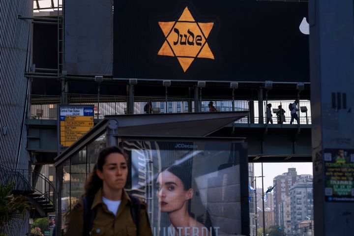 People walk along a bridge lit with a billboard showing a yellow Star of David that reads "Jude", Jew in German, resembling the one Jews were forced to wear in Nazi Germany, during the annual Holocaust Remembrance Day in Ramat Gan, Israel, Thursday, April 28, 2022. (AP Photo/Oded Balilty)