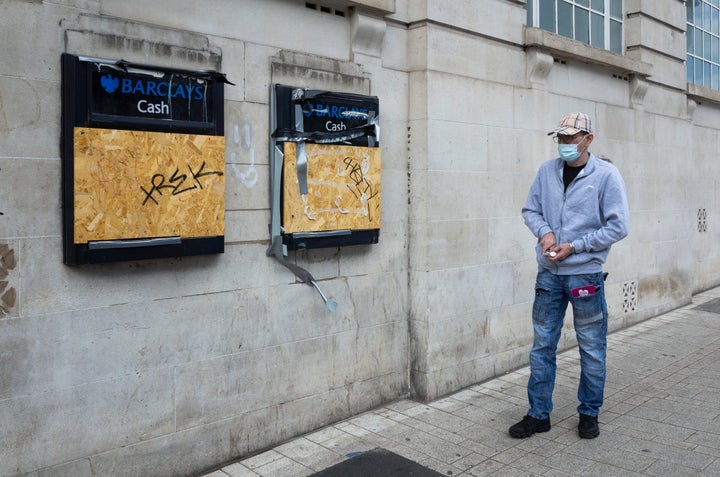 A man wearing a face mask to protect against Covid-19 inspects two Barclays cash points, both boarded up and vandalised by graffiti.