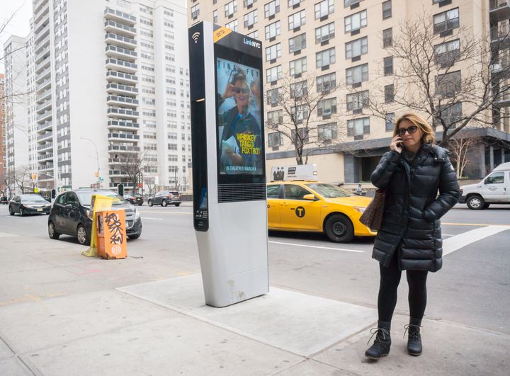 A woman stands near a LinkNYC kiosk in New York City. These kiosks provide free Wi-Fi, phone charging and phone calls. The system is supported by advertising running on the sides of the kiosks.