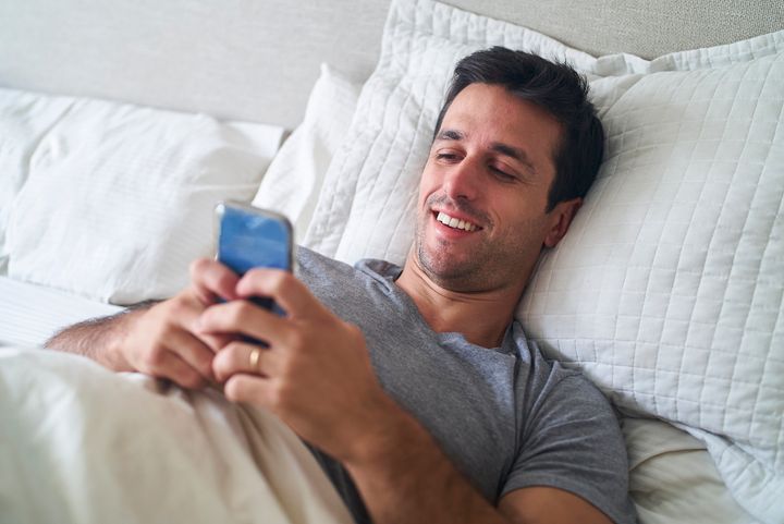 high angle shot of hispanic adult man lying in bed while using smart phone during daytime