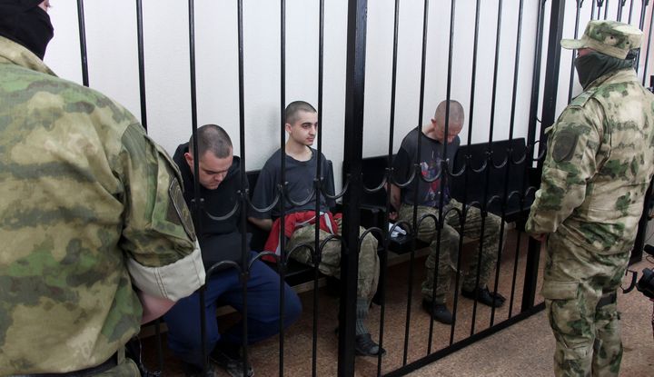 Two British citizens Aiden Aslin, left, and Shaun Pinner, right, and Moroccan Saaudun Brahim, center, sit behind bars in a courtroom in Donetsk.