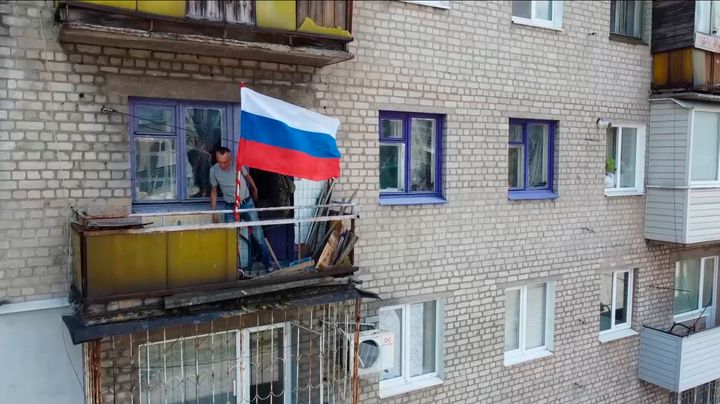 In this handout photo taken from video released by Russian Defense Ministry Press Service on July 4, 2022, a man sets a Russian national flag on a balcony of a residential building in Lysychansk.