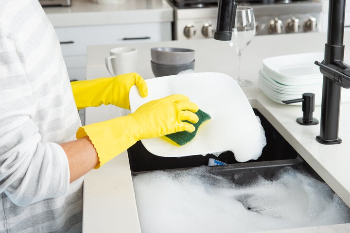 Unrecognizable hand with yellow cleaning glove scrubbing dishes close up in kitchen.