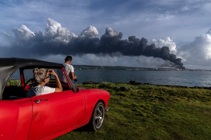 People watch a huge plume of smoke rise from the Matanzas supertanker base, as firefighters work to douse a fire that started during a thunderstorm the night before, in Matanzas, Cuba, Sunday, Aug. 7, 2022. Cuban authorities say lightning struck a crude oil storage tank at the base, sparking a fire that sparked four explosions that injured more than 121 people, one person dead and 17 missing. (AP Photo/Ramon Espinosa)