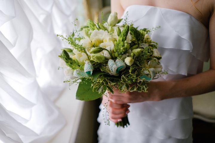 A beautiful bouquet with white flowers in the hands of a girl or a woman. The bride in a luxurious expensive elegant dress holds a wedding bouquet of roses and green leaves in her hands, indoors.