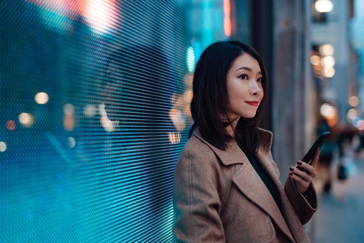 A young woman holds a phone while waiting on a city street at night