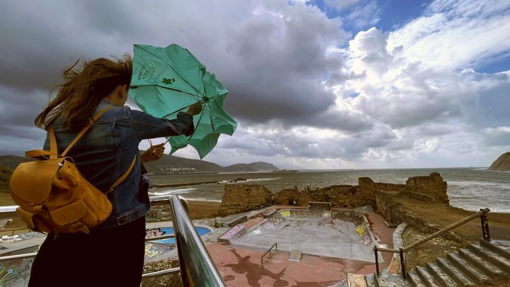 Una mujer trata de abrir su paraguas frente a la playa de Arrigunaga en Getxo (Bizkaia), este miércoles, en el que el calor ha dado paso a fuertes lluvias y rachas de viento intenso.