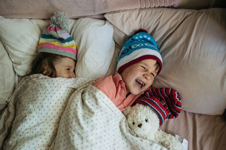 Siblings sleeping in their bed wearing winter hats and holding a teddy bear. 