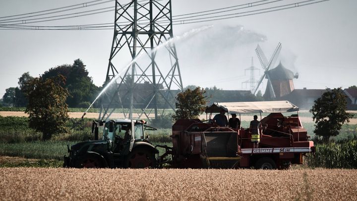 20 Ιουλίου 2022, Κάτω Σαξονία, Wendeburg: Ένα χωράφι κοντά στον μύλο του Zweidorf (πίσω) ποτίζεται ενώ ένας αγρότης μαζεύει πατάτες με τους βοηθούς του.