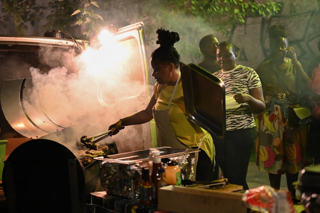 A woman grills jerk chicken on All Saints Road as the Mangrove Steelband rehearses.