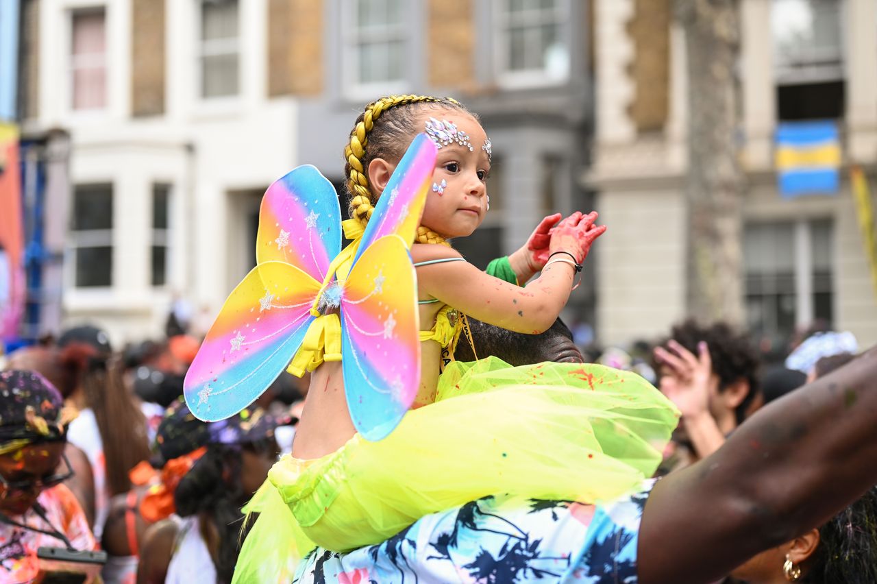 A young girl perched on a man's shoulders wears bright butterfly wings and observes the first day of the Notting Hill Carnival parade. The first Carnival parade is described as family-friendly.