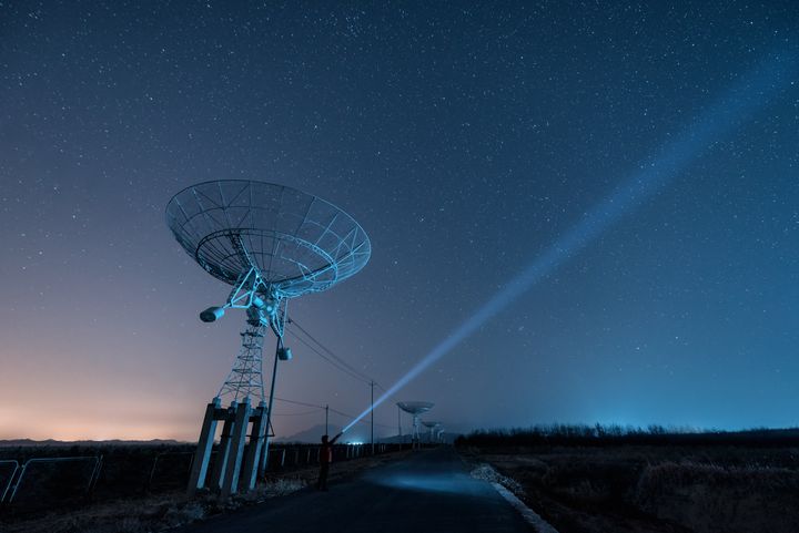 Ένας άνδρας στέκεται κάτω από ένα ραδιοτηλεσκόπιο τη νύχτα, κρατώντας έναν φακό, ενώ μια δέσμη φωτός εκτοξεύεται προς τον έναστρο ουρανό.