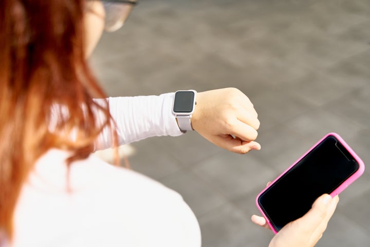 Teenager with long hair wear white shirt and she looks at her smartwatch while holding a cell phone with other hand