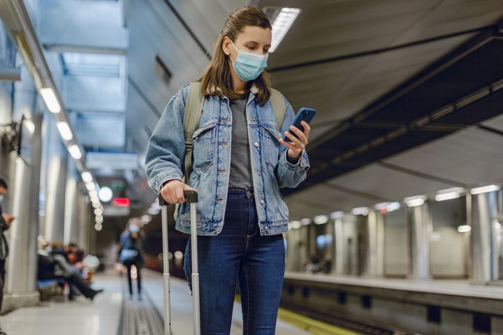 Photo of a young woman with protective face mask holding mobile phone while waiting for the train