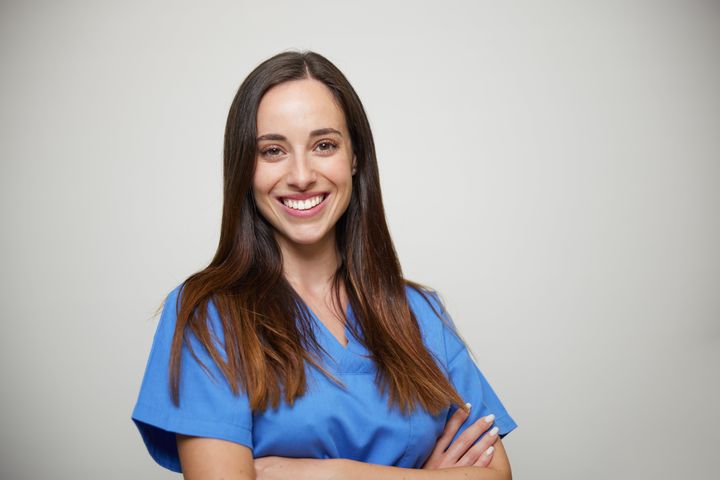 A woman in front of a plain grey background is wearing medical blue scrubs.