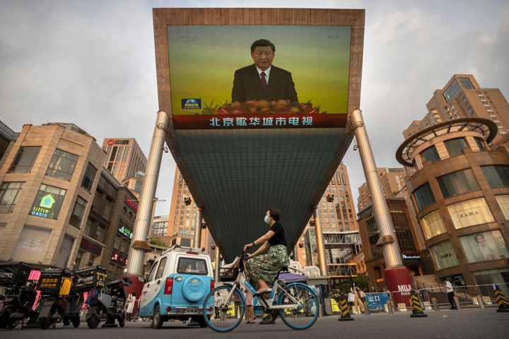 Μια γυναίκα που φοράει μάσκα προσώπου οδηγεί ένα ποδήλατο στο Πεκίνο κάτω από μια μεγάλη οθόνη που εμφανίζει ειδησεογραφική κάλυψη της κινεζικής κρατικής τηλεόρασης της επίσκεψης του προέδρου Xi Jinping στο Χονγκ Κονγκ για τον εορτασμό της 25ης επετείου από την επιστροφή του Χονγκ Κονγκ στην Κίνα την 1η Ιουλίου 2022. (AP Photo/Mark Schiefelbein)
