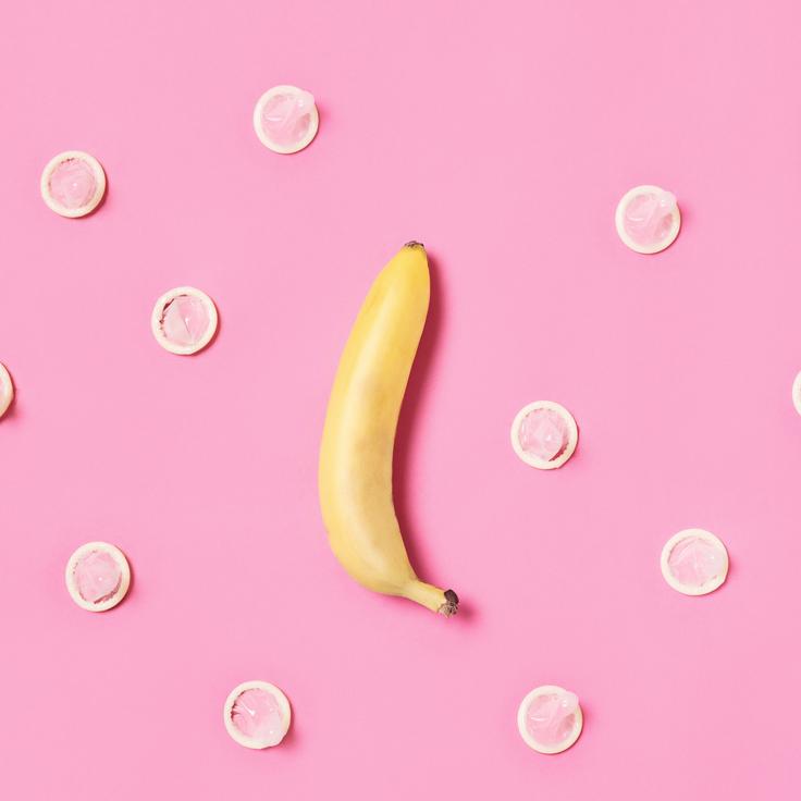 A yellow banana on a pink background surrounded by latex condoms in a random pattern. Concept to illustrate safe sex and male contraception. Shot with a with a top down view