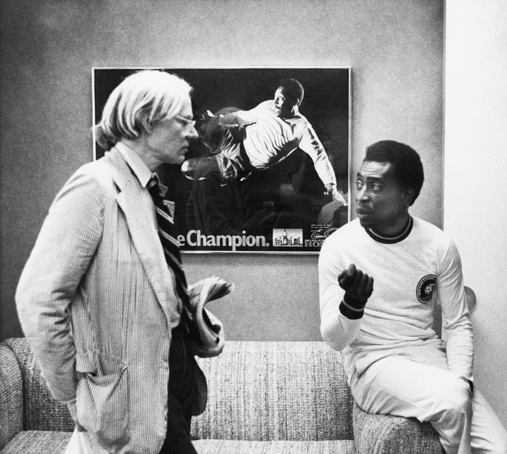 Ο καλλιτέχνης Andy Warhol, αριστερά, συνομιλεί με τον σούπερ σταρ του ποδοσφαίρου Pele για ένα πορτρέτο που του κάνει, 26 Ιουλίου 1977, Νέα Υόρκη. Στο βάθος διακρίνεται μια αφίσα με μια φωτογραφία του Πελέ εν δράσει.