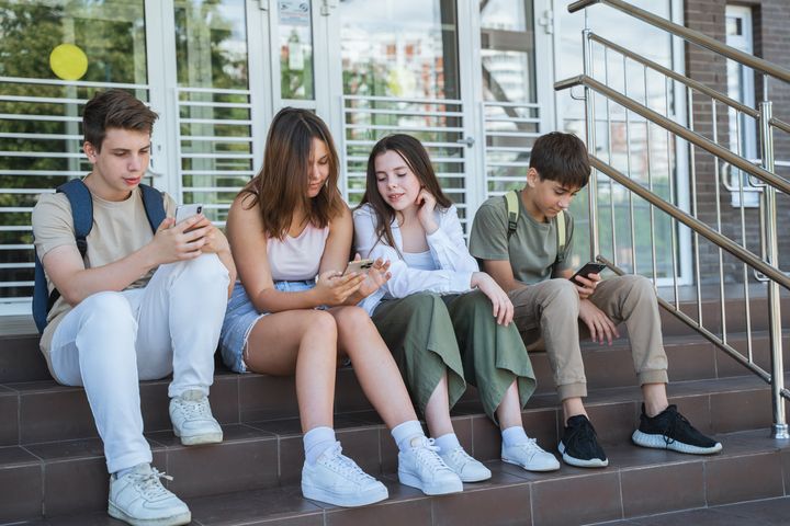 Group of teenagers surfing internet, using social media, sit on steps near school