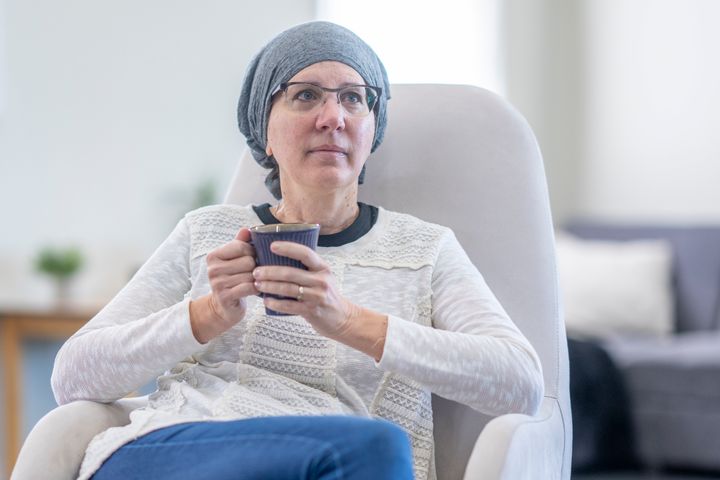 A middle aged woman sits in a high-back chair as she sips a cup of tea. She is dressed casually and has a headscarf on to keep her warm between cancer treatments.