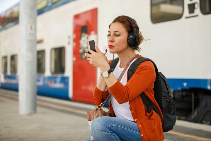 Copy space shot of mid adult woman refreshing her make up at the the railroad station. She is crouching, holding a small hand mirror and applying lipstick before boarding the train.