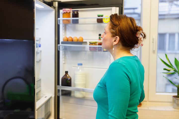 Woman standing by the open fridge and looking inside for food at meal time
