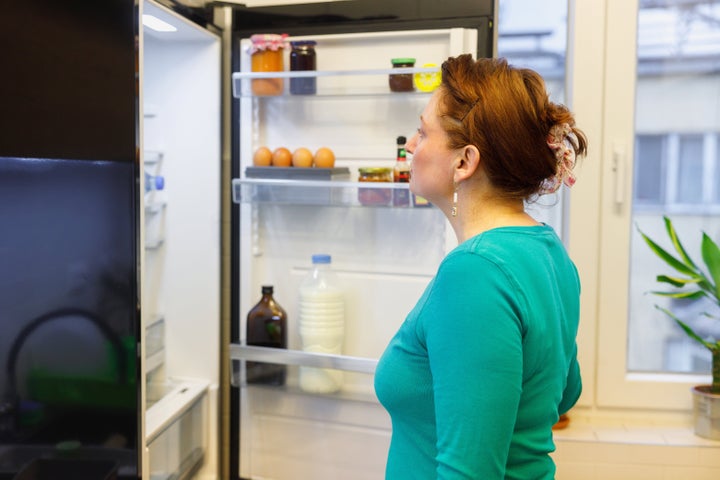 Woman standing by the open fridge and looking inside for food at meal time