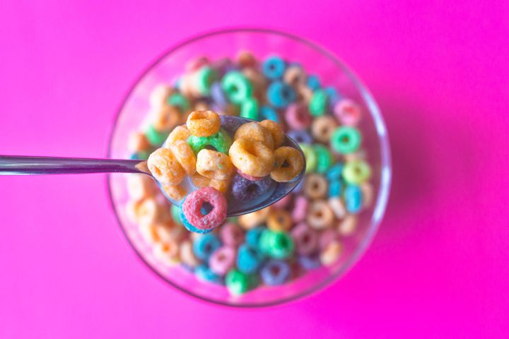 Colorful breakfast cereal with milk fills spoon in foreground with cereal in glass bowl on pink background in out-of-focus background