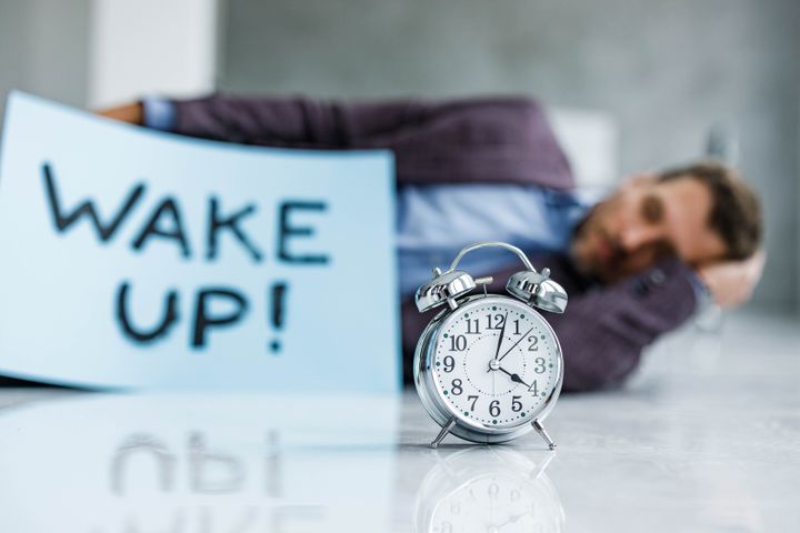 Close up of an alarm clock on the floor with a businessman in the background sleeping. Copy space.