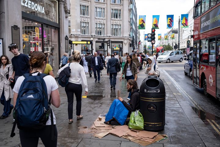 Shoppers and visitors out on Oxford Street walk past a homeless man sitting against a rubbish bin while asking for money.